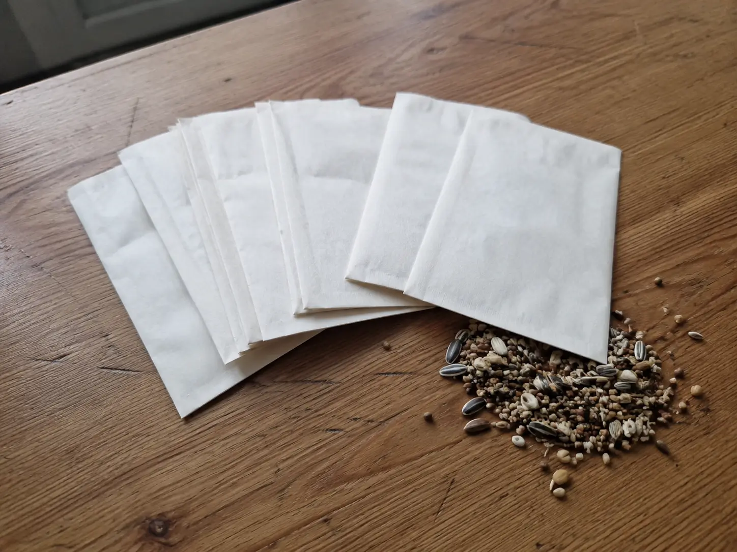 Blank seed packets fanned out on a wooden table with a small pile of mixed seeds beside them